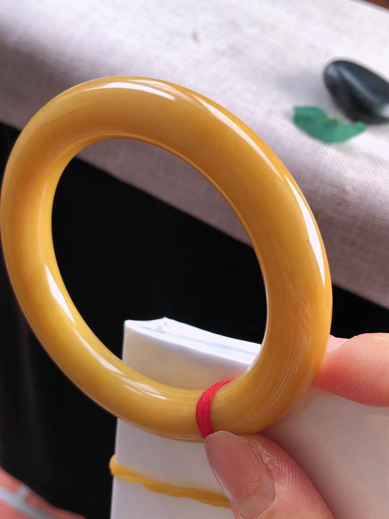 K&ouml;nnte beinhalten: Ein poliertes, bernsteinfarbenes Armreif-Armband. Das kreisf&ouml;rmige Armband wird von einer Hand gehalten. Das Armband hat eine glatte, gl&auml;nzende Oberfl&auml;che. Der Hintergrund ist verschwommen, mit Andeutungen von Wei&szlig; und Schwarz.