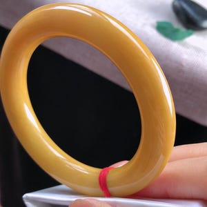 K&ouml;nnte beinhalten: Ein glattes, bernsteinfarbenes Armreif-Armband. Das kreisf&ouml;rmige Armband hat eine einfarbige, gl&auml;nzende Oberfl&auml;che. Ein kleiner roter Faden ist um einen Teil des Armbands gewickelt. Das Armband wird &uuml;ber einer wei&szlig;en Oberfl&auml;che gehalten.