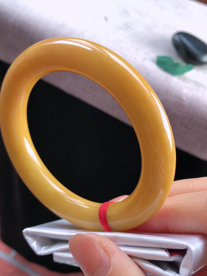 K&ouml;nnte beinhalten: Ein glattes, bernsteinfarbenes Armreif-Armband. Das kreisf&ouml;rmige Armband hat eine einfarbige, gl&auml;nzende Oberfl&auml;che. Ein kleiner roter Faden ist um einen Teil des Armbands gewickelt. Das Armband wird &uuml;ber einer wei&szlig;en Oberfl&auml;che gehalten.