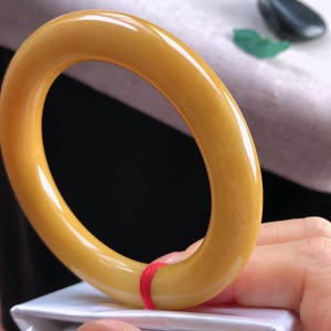 K&ouml;nnte beinhalten: Ein gl&auml;nzendes, bernsteinfarbenes Armreif-Armband. Das kreisf&ouml;rmige Armband ist ein durchgehendes, glattes Band. Ein roter Faden ist um einen Teil des Armbands gewickelt. Der Hintergrund umfasst eine wei&szlig;e Oberfl&auml;che und einen schwarzen Hintergrund.