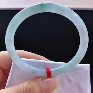 May include: A pale green and white jade bangle bracelet. The translucent stone has a polished surface, with a deeper green section. The bracelet is held against a white fabric, and a red ribbon is visible. The bracelet is a circular shape.