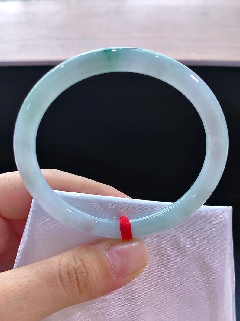 May include: A pale green and white jade bangle bracelet. The translucent stone has a polished surface, with a deeper green section. The bracelet is held against a white fabric, and a red ribbon is visible. The bracelet is a circular shape.