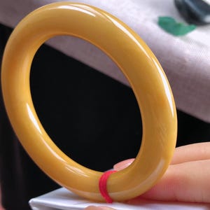 K&ouml;nnte beinhalten: Ein glattes, goldgelbes Armreif-Armband. Das Armband hat eine runde Form und eine polierte Oberfl&auml;che. Ein kleiner roter Faden ist um einen Teil des Armbands gewickelt. Der Hintergrund ist eine Mischung aus Wei&szlig; und Schwarz.