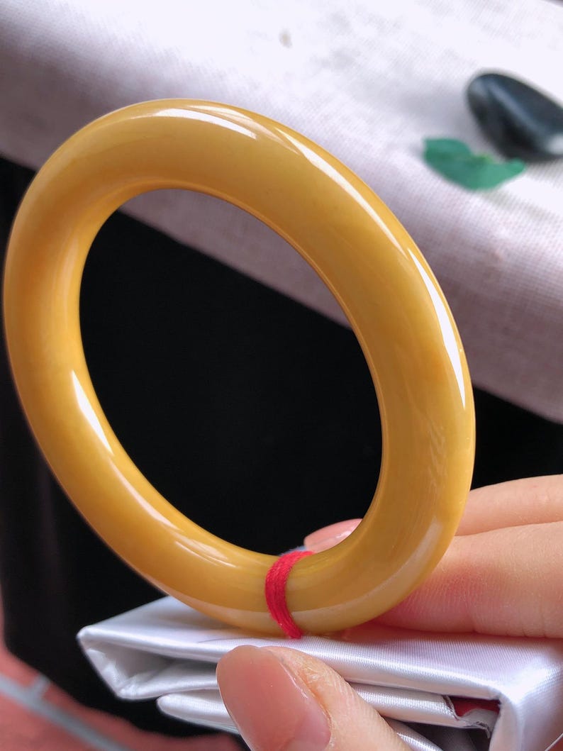 K&ouml;nnte beinhalten: Ein glattes, goldgelbes Armreif-Armband. Das Armband hat eine runde Form und eine polierte Oberfl&auml;che. Ein kleiner roter Faden ist um einen Teil des Armbands gewickelt. Der Hintergrund ist eine Mischung aus Wei&szlig; und Schwarz.