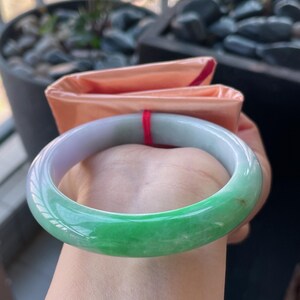 K&ouml;nnte beinhalten: Ein Jade-Armreif mit einem Farbverlauf von hellem Lila zu leuchtendem Gr&uuml;n. Die polierte Oberfl&auml;che reflektiert das Licht und hebt die nat&uuml;rlichen Variationen des Steins hervor. Ein klassisches Schmuckst&uuml;ck.