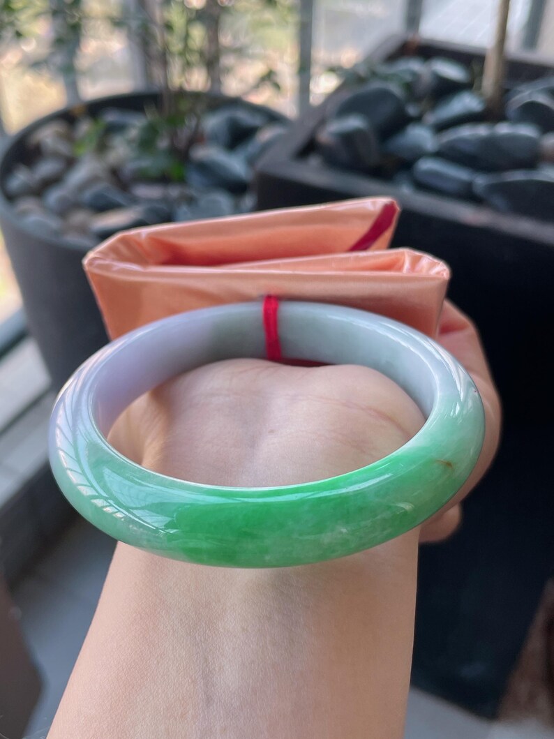 K&ouml;nnte beinhalten: Ein Jade-Armreif mit einem Farbverlauf von hellem Lila zu leuchtendem Gr&uuml;n. Die polierte Oberfl&auml;che reflektiert das Licht und hebt die nat&uuml;rlichen Variationen des Steins hervor. Ein klassisches Schmuckst&uuml;ck.