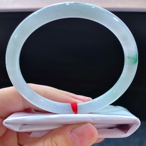 May include: A pale green jadeite bangle. The translucent bracelet has a smooth, polished finish with a touch of green. The bracelet is resting on a white package with a red ribbon.