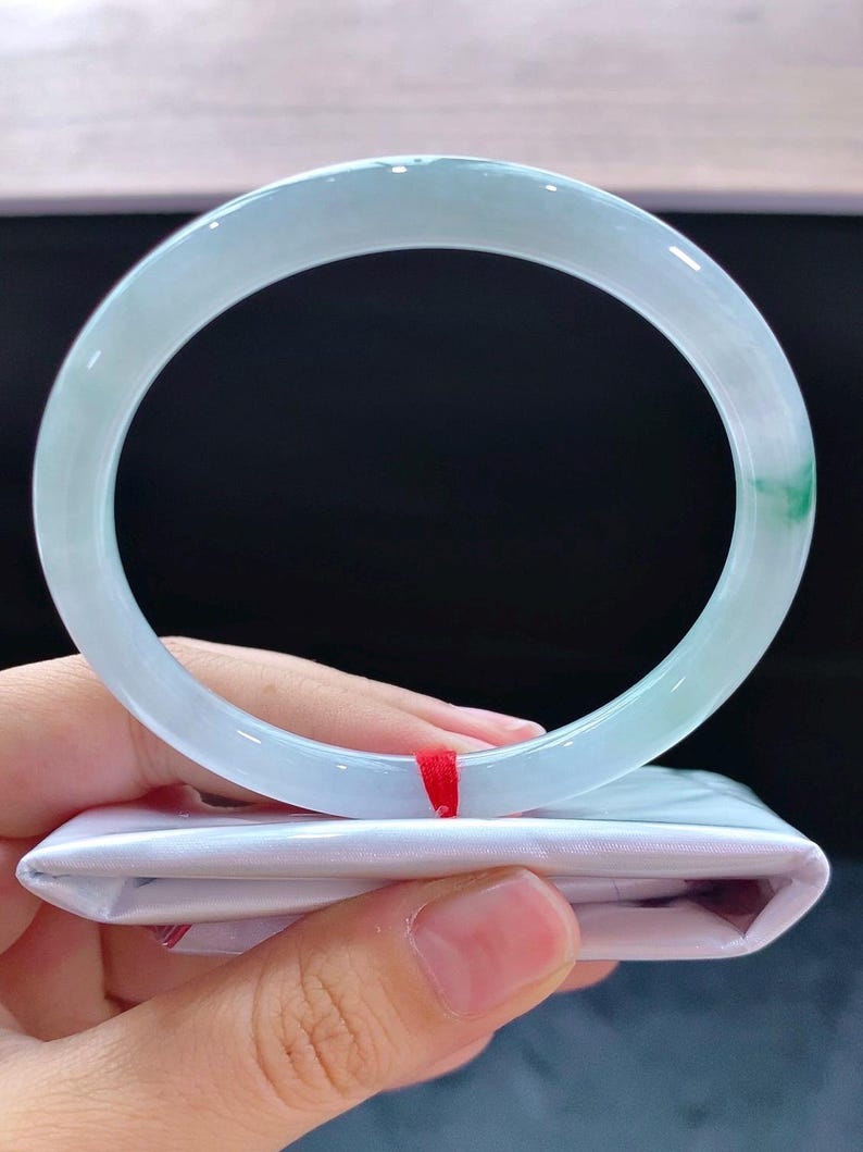 May include: A pale green jadeite bangle. The translucent bracelet has a smooth, polished finish with a touch of green. The bracelet is resting on a white package with a red ribbon.