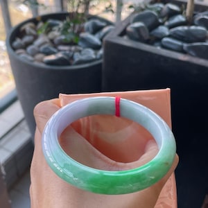 K&ouml;nnte beinhalten: Ein Jade-Armreif mit einem Farbverlauf, der von hellem Lavendel zu lebendigem Gr&uuml;n &uuml;bergeht. Die glatte, polierte Oberfl&auml;che reflektiert das Licht und zeigt die nat&uuml;rlichen Variationen des Steins. Ein roter Faden ist am Armreif befestigt.