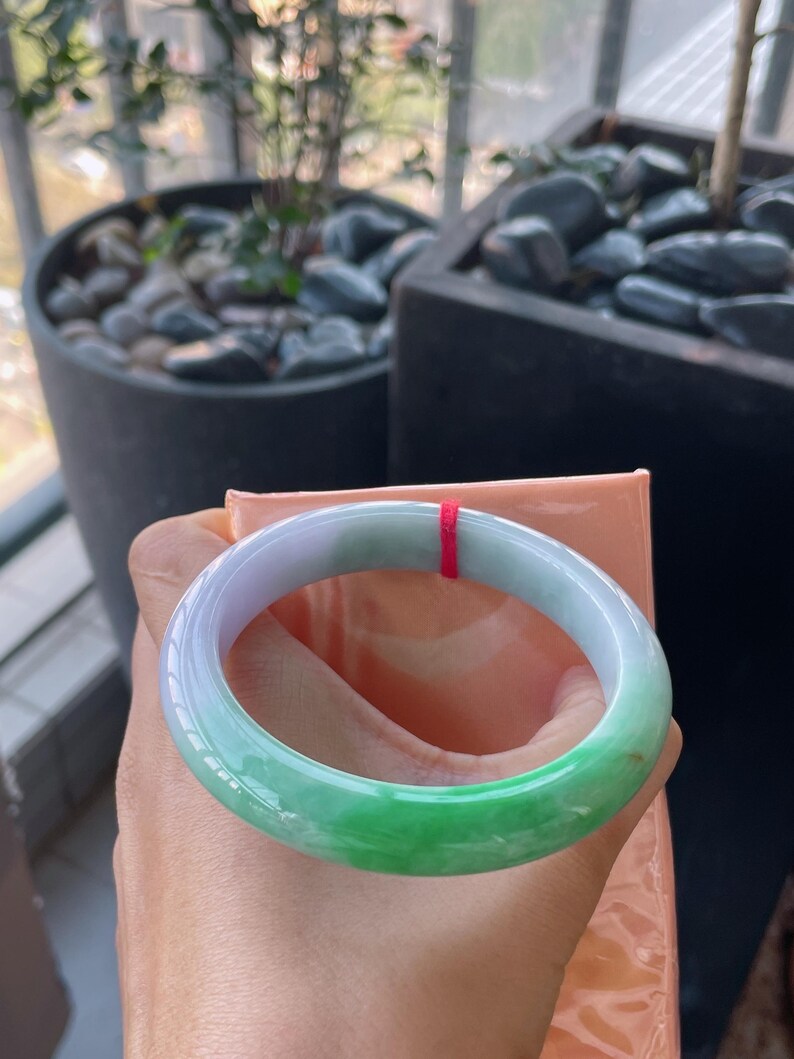 K&ouml;nnte beinhalten: Ein Jade-Armreif mit einem Farbverlauf, der von hellem Lavendel zu lebendigem Gr&uuml;n &uuml;bergeht. Die glatte, polierte Oberfl&auml;che reflektiert das Licht und zeigt die nat&uuml;rlichen Variationen des Steins. Ein roter Faden ist am Armreif befestigt.