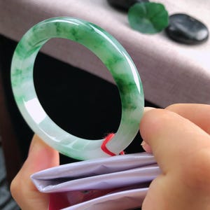 May include: A jade bangle with a translucent green and white pattern. The polished stone is held in a hand, highlighting its smooth surface. The bangle's design is a classic, circular shape. Other jade stones are in the background.