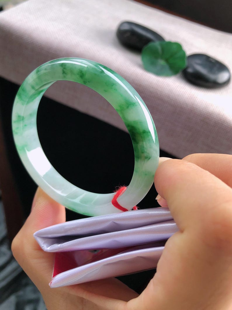 May include: A jade bangle with a translucent green and white pattern. The polished stone is held in a hand, highlighting its smooth surface. The bangle's design is a classic, circular shape. Other jade stones are in the background.