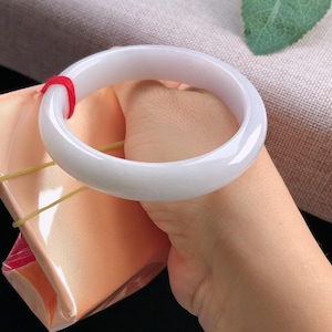 May include: A smooth, white jade bangle bracelet. The bracelet is held in a hand, with a red string visible. The background includes a pink fabric and a green leaf.