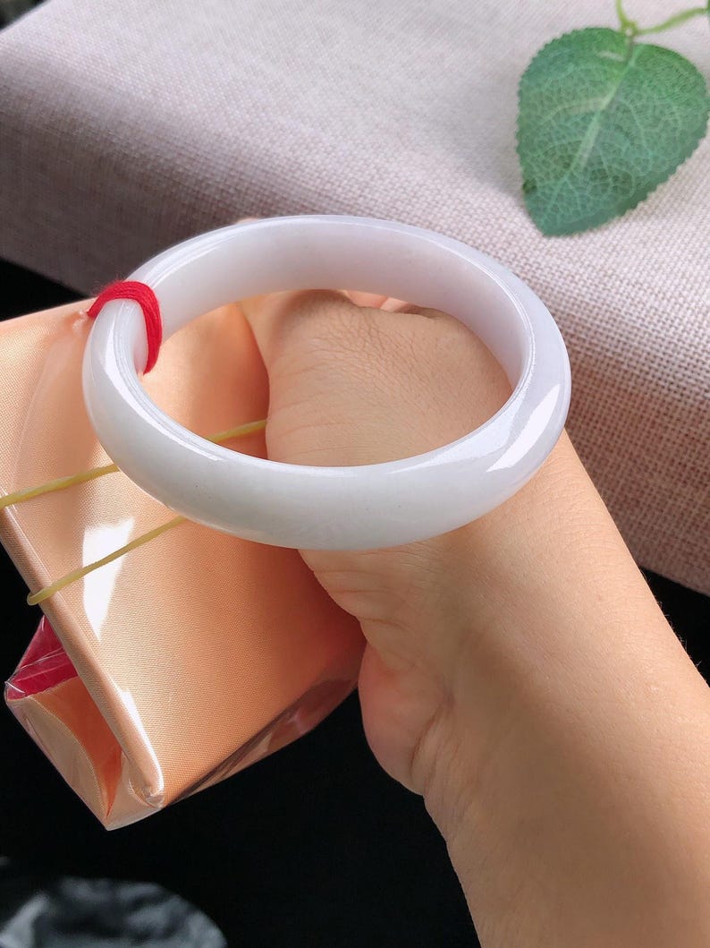 May include: A smooth, white jade bangle bracelet. The bracelet is held in a hand, with a red string visible. The background includes a pink fabric and a green leaf.