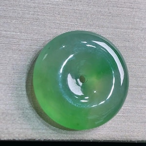 Könnte beinhalten: Runder, durchscheinender Jade-Anhänger in Grün mit zentralem Loch. Die polierte, reflektierende Oberfläche zeigt eine glatte Textur und einen subtilen Farbverlauf. Ein Schmuckstück aus Jade.