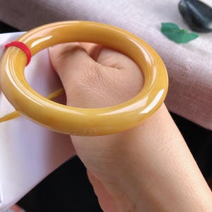 K&ouml;nnte beinhalten: Ein glattes, gelb get&ouml;ntes Armreif-Armband. Das Armband ist ein massives, kreisf&ouml;rmiges Band mit polierter Oberfl&auml;che. Ein roter Faden ist um einen Teil des Armbands gewickelt. Das Armband wird von einer Person gehalten.