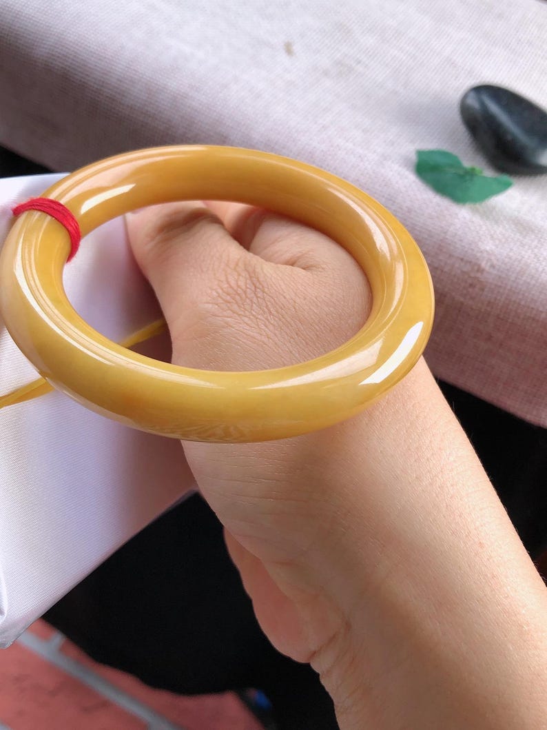 K&ouml;nnte beinhalten: Ein glattes, gelb get&ouml;ntes Armreif-Armband. Das Armband ist ein massives, kreisf&ouml;rmiges Band mit polierter Oberfl&auml;che. Ein roter Faden ist um einen Teil des Armbands gewickelt. Das Armband wird von einer Person gehalten.