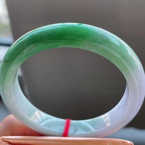 K&ouml;nnte beinhalten: Ein Jade-Armreif mit glatter, polierter Oberfl&auml;che. Das Armband geht von einem leuchtenden Gr&uuml;n in ein durchscheinendes Wei&szlig; &uuml;ber. Der Jade ist eine solide, kreisf&ouml;rmige Form und liegt auf einem gefalteten orangefarbenen Papier.