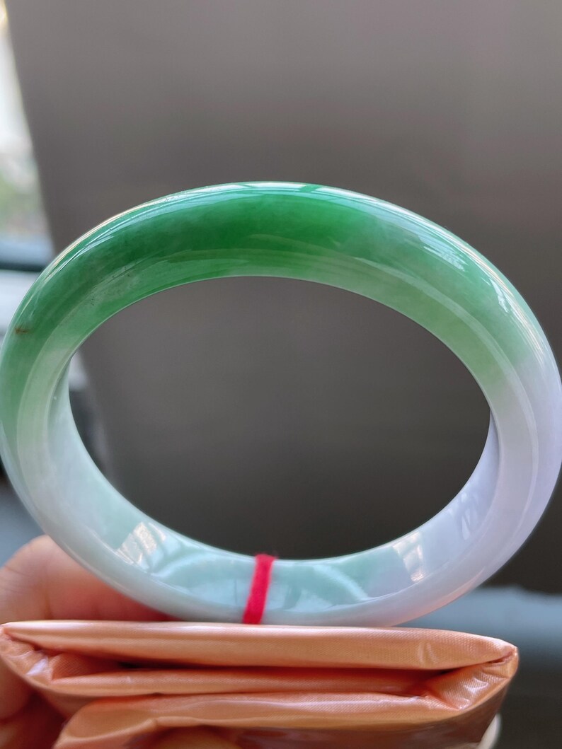 K&ouml;nnte beinhalten: Ein Jade-Armreif mit glatter, polierter Oberfl&auml;che. Das Armband geht von einem leuchtenden Gr&uuml;n in ein durchscheinendes Wei&szlig; &uuml;ber. Der Jade ist eine solide, kreisf&ouml;rmige Form und liegt auf einem gefalteten orangefarbenen Papier.