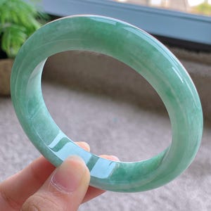 May include: A translucent, jade green bangle bracelet. The smooth, polished surface reflects light, showcasing the natural variations in the stone. The bracelet is a solid, circular band, ideal for everyday wear.