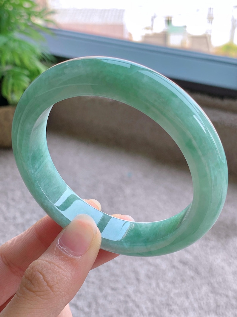 May include: A translucent, jade green bangle bracelet. The smooth, polished surface reflects light, showcasing the natural variations in the stone. The bracelet is a solid, circular band, ideal for everyday wear.