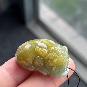 Peut inclure: Pendentif en jade sculpt&eacute; en forme de cr&eacute;ature mythique. Le pendentif est un m&eacute;lange de teintes vertes et jaunes, avec des d&eacute;tails complexes sur la t&ecirc;te et le corps. Un cordon noir est attach&eacute; pour le porter.