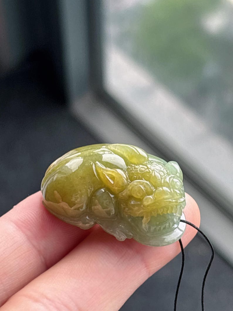 Peut inclure: Pendentif en jade sculpt&eacute; en forme de cr&eacute;ature mythique. Le pendentif est un m&eacute;lange de teintes vertes et jaunes, avec des d&eacute;tails complexes sur la t&ecirc;te et le corps. Un cordon noir est attach&eacute; pour le porter.