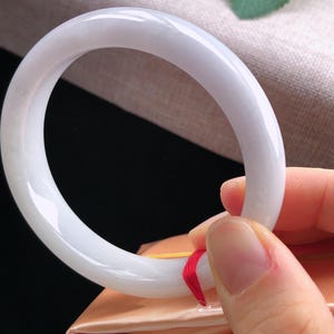 May include: A smooth, white jade bangle bracelet. The circular bracelet is held up by a hand, showcasing its polished surface and subtle translucence. The jade is a soft, creamy white color, reflecting light.