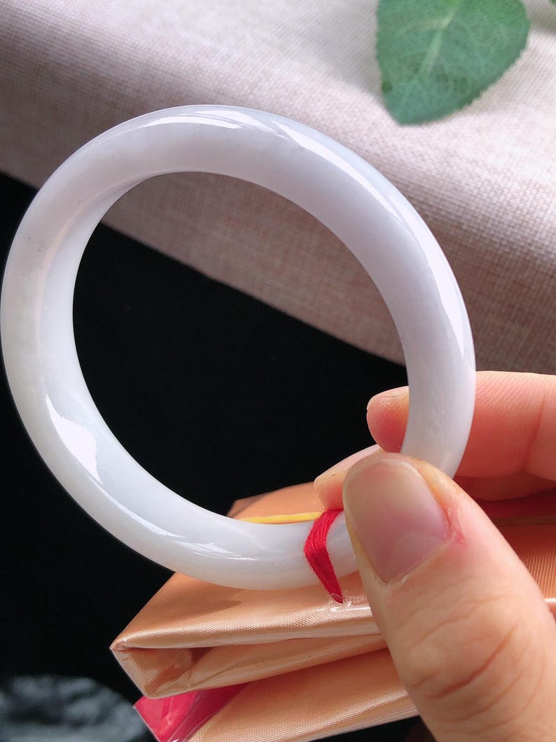 May include: A smooth, white jade bangle bracelet. The circular bracelet is held up by a hand, showcasing its polished surface and subtle translucence. The jade is a soft, creamy white color, reflecting light.