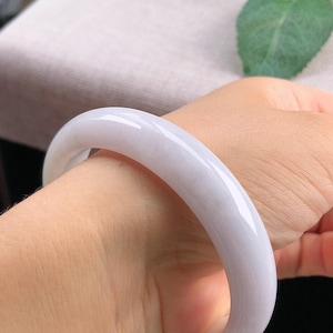 May include: A smooth, pale lavender jade bangle bracelet. The polished stone has a rounded shape and is worn on a wrist. The bracelet's color is a soft, muted purple, and it reflects light, highlighting its glossy surface.