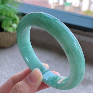 May include: A light green jade bangle bracelet. The smooth, polished surface of the bracelet reflects light, showcasing its translucent quality. The jade is a soft, muted green color.
