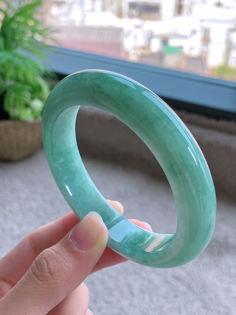 May include: A light green jade bangle bracelet. The smooth, polished surface of the bracelet reflects light, showcasing its translucent quality. The jade is a soft, muted green color.