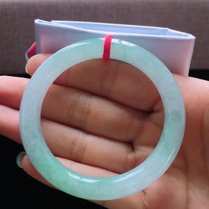 Op de afbeelding: Een doorschijnende jade armband met een verloop van lichtgroen naar wit. Het gladde, gepolijste oppervlak reflecteert licht en toont de natuurlijke kleurvariaties. Een rood draadje is aan de armband bevestigd.