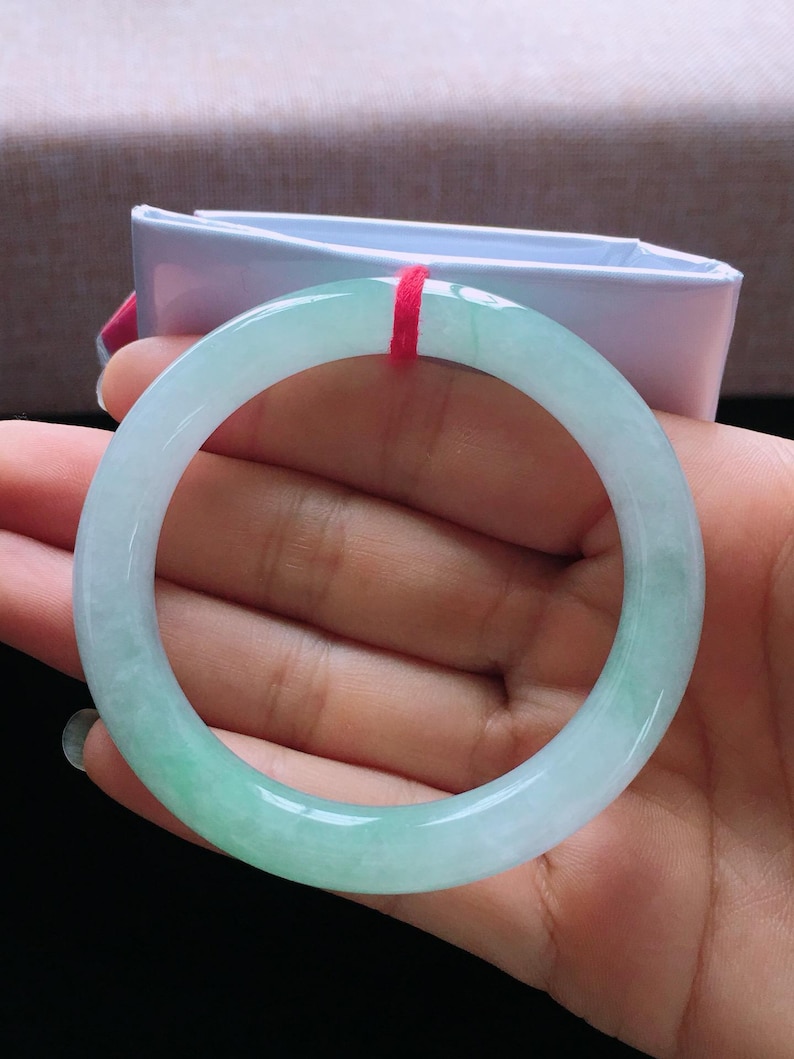 Op de afbeelding: Een doorschijnende jade armband met een verloop van lichtgroen naar wit. Het gladde, gepolijste oppervlak reflecteert licht en toont de natuurlijke kleurvariaties. Een rood draadje is aan de armband bevestigd.