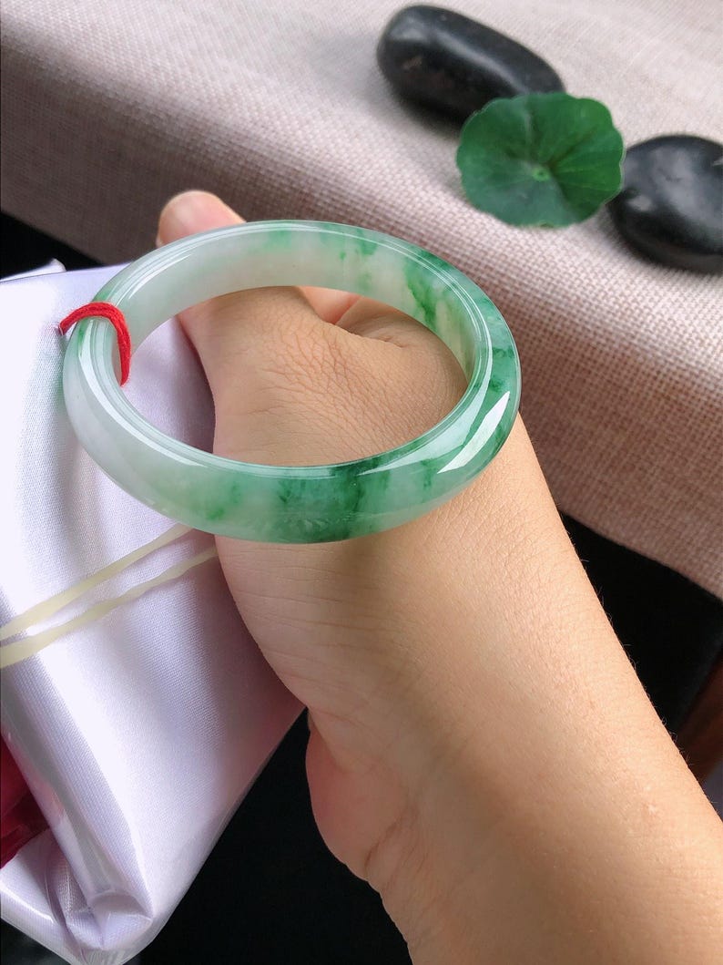 May include: A jade bangle bracelet with a translucent green and white colour gradient. The polished stone is smooth and circular, with a red string tied around it. The bracelet is held in a hand.