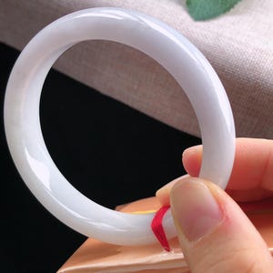 May include: A smooth, white jade bangle bracelet. The polished surface reflects light, showcasing a subtle, translucent quality. The bracelet is held by a hand, revealing a red ribbon attached to the bracelet.