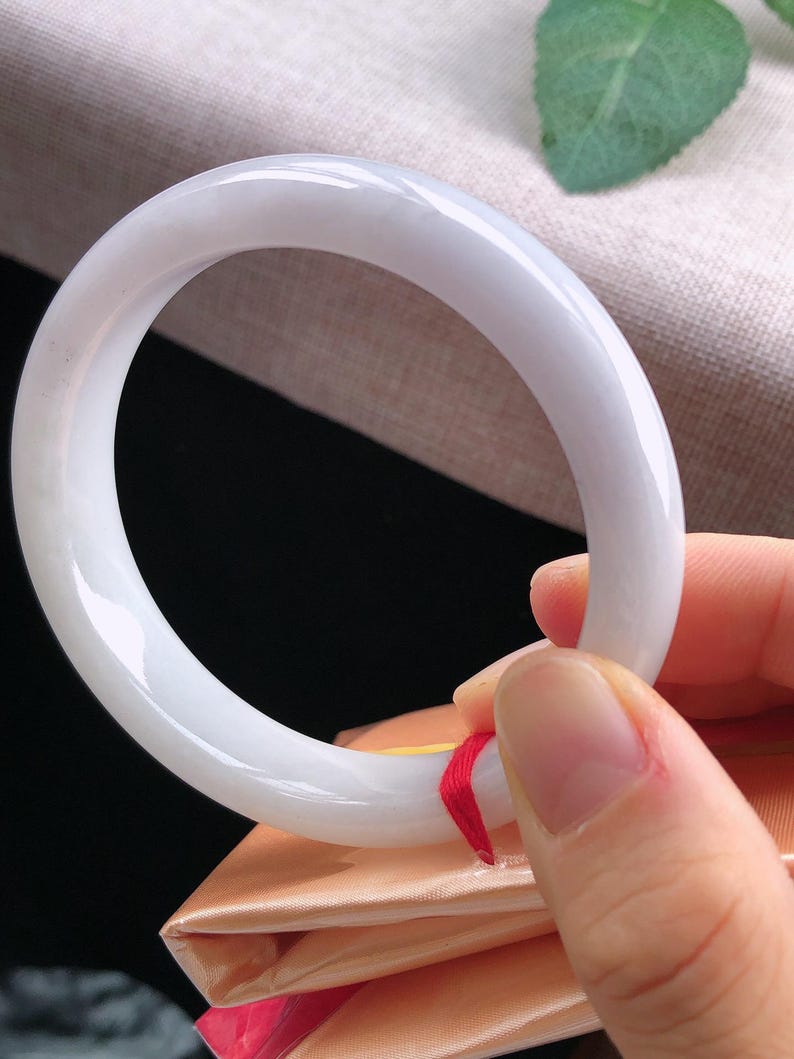 May include: A smooth, white jade bangle bracelet. The polished surface reflects light, showcasing a subtle, translucent quality. The bracelet is held by a hand, revealing a red ribbon attached to the bracelet.