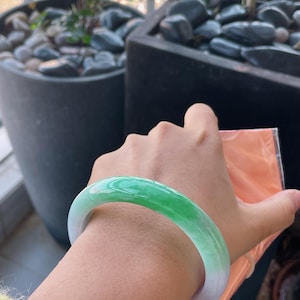 K&ouml;nnte beinhalten: Ein Jade-Armreif mit einem Farbverlauf aus Gr&uuml;n- und Wei&szlig;t&ouml;nen. Die polierte Oberfl&auml;che reflektiert das Licht und hebt die nat&uuml;rlichen Farbvariationen hervor. Das Armband hat eine kreisf&ouml;rmige Form und ist zum Tragen am Handgelenk bestimmt.