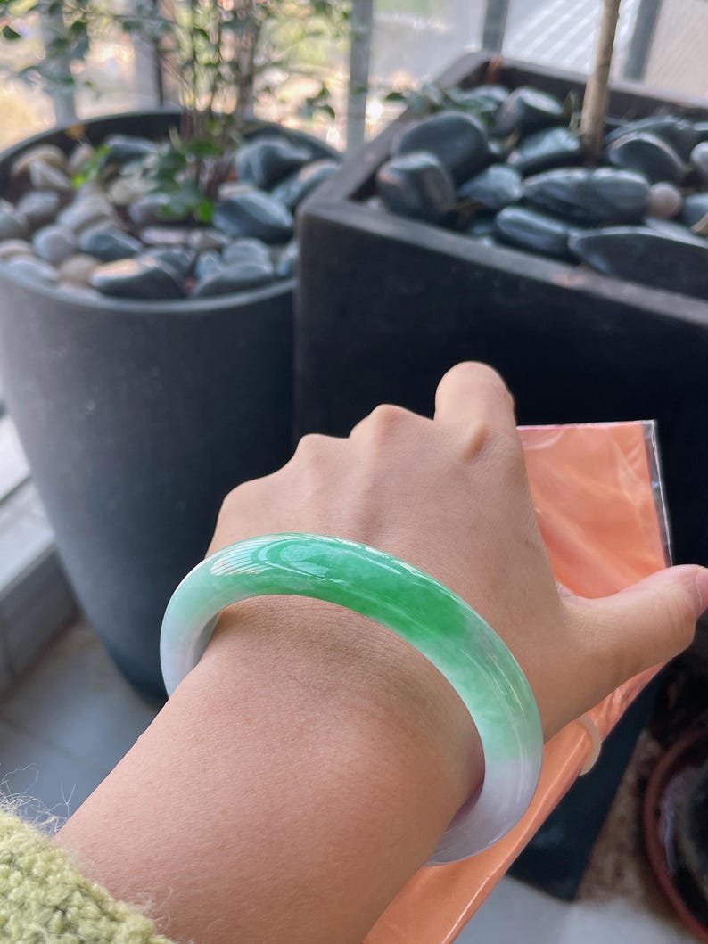 K&ouml;nnte beinhalten: Ein Jade-Armreif mit einem Farbverlauf aus Gr&uuml;n- und Wei&szlig;t&ouml;nen. Die polierte Oberfl&auml;che reflektiert das Licht und hebt die nat&uuml;rlichen Farbvariationen hervor. Das Armband hat eine kreisf&ouml;rmige Form und ist zum Tragen am Handgelenk bestimmt.