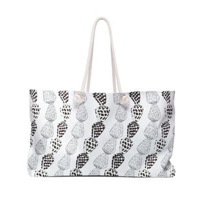 May include: White tote bag with a repeating black and white seashell pattern. The bag has a rectangular shape, a white rope handle, and a small white pom-pom detail. The bag is designed for carrying items.