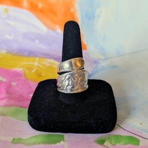 May include: A silver spoon ring with a floral design on the lower band and a smooth, curved upper band. The ring is displayed on a black velvet ring holder against a colorful background.