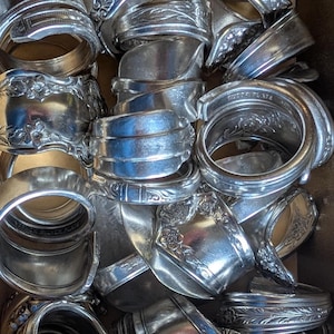 May include: A collection of silver-colored rings, crafted from repurposed silverware. The rings display various floral and ornate patterns, with some bearing stamped markings. The rings are arranged in a close-up, top-down view.