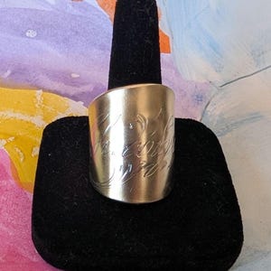 May include: A silver-colored ring with engraved cursive script. The ring is wide and has a smooth, polished surface. The ring is displayed on a black velvet ring holder against a colorful background.