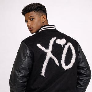 May include: Black varsity jacket with white "XO" graphic on the back. The jacket has black leather sleeves, a striped collar, and a striped hem. The model is wearing blue jeans.