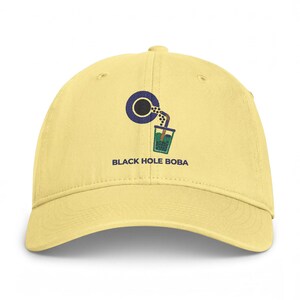 May include: A pale yellow baseball cap featuring an embroidered graphic of a dark blue black hole pouring boba pearls into a green boba tea cup with a straw. The text BLACK HOLE BOBA is embroidered in dark blue below the graphic design.