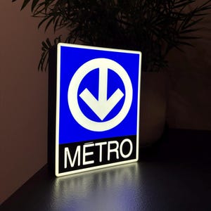 Può includere: Un cartello della metropolitana blu e bianco con una freccia bianca rivolta verso il basso all'interno di un cerchio. La parola "METRO" è scritta in bianco su sfondo nero. Il cartello è illuminato, probabilmente un display luminoso, per i trasporti pubblici.