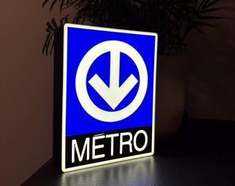 May include: A blue and white Metro sign with a downward-pointing arrow inside a white circle. The word "METRO" is displayed in white letters on a black background. The sign is illuminated, suggesting it is a light-up sign.