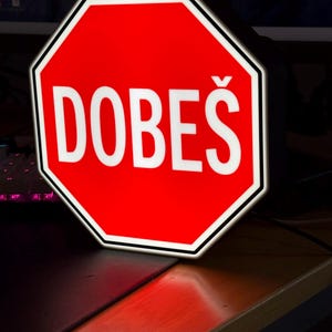 Op de afbeelding: Een rood, achthoekig bord met het woord "DOBEŠ" in witte letters. Het bord heeft een witte rand en is verlicht, waarschijnlijk een decoratief of nieuwigheidsobject. Het bord staat op een houten oppervlak.