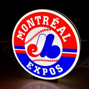 Puede incluir: Un letrero circular iluminado con el logotipo del equipo de béisbol Montreal Expos. El diseño incluye el emblema estilizado 'eb' en rojo, blanco y azul, centrado dentro de un borde circular que contiene el texto 'MONTRÉAL' y 'EXPOS' en blanco.