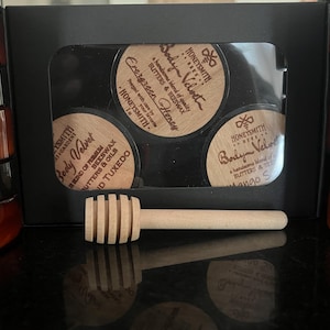 Könnte beinhalten: Ein schwarzes Set mit drei Gläsern Körperbutter mit Holzdeckeln und einem hölzernen Honiglöffel. Die Gläser sind mit "Body Velvet" und "Evergreen Honey" mit dem Markennamen "Honeysmith" beschriftet.